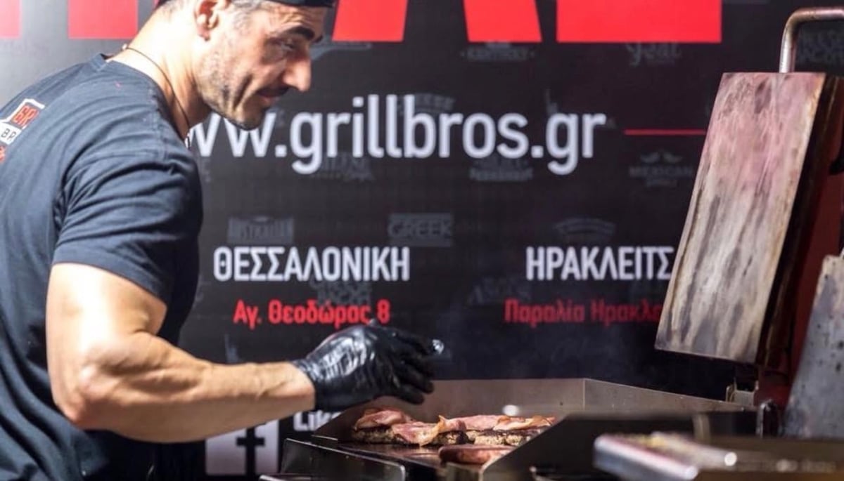 Man in blue shirt grilling meat at a restaurant counter with Greek signage